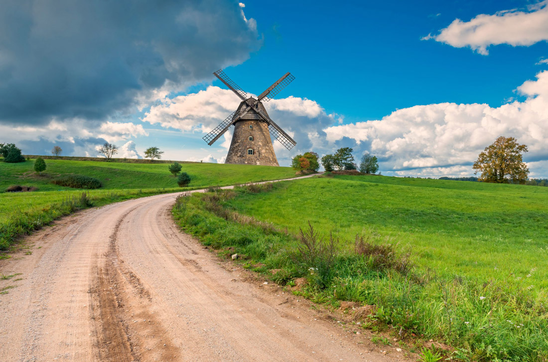 Topic is Travel Destination to Latvia. Countryside gravel road among spring fields and leading to an old windmill.