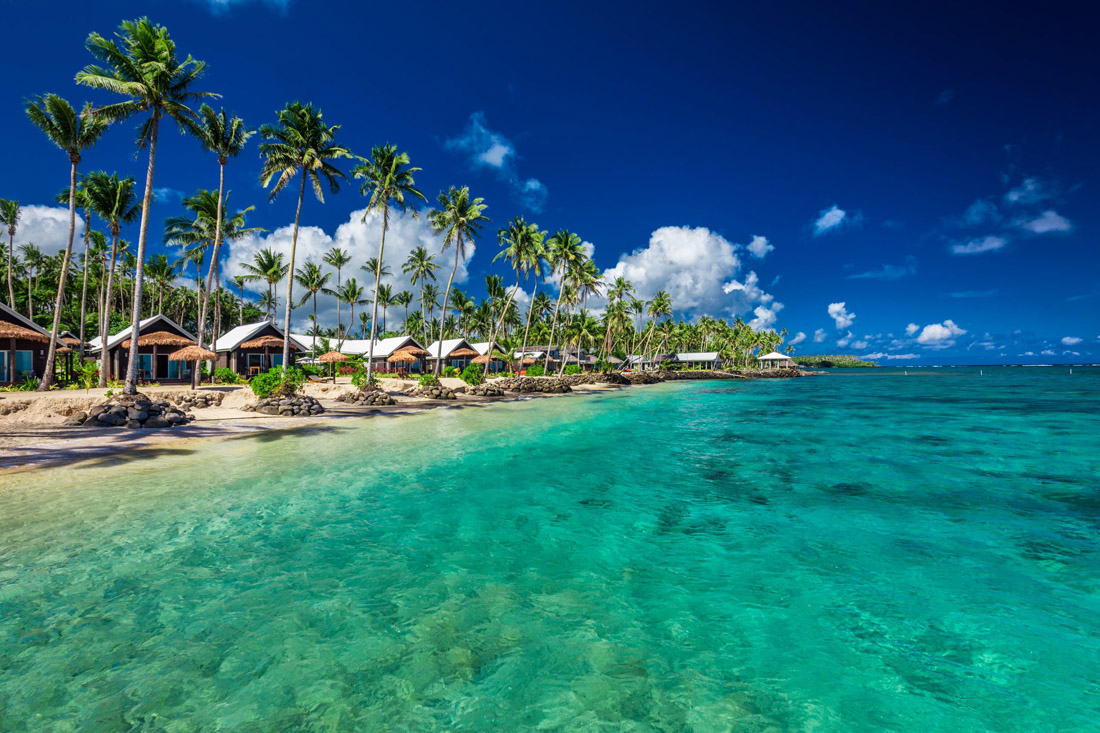 Topic is Travel Destination to Samoa. Tropical beach with coconut palm trees and villas in scenic Samoa.
