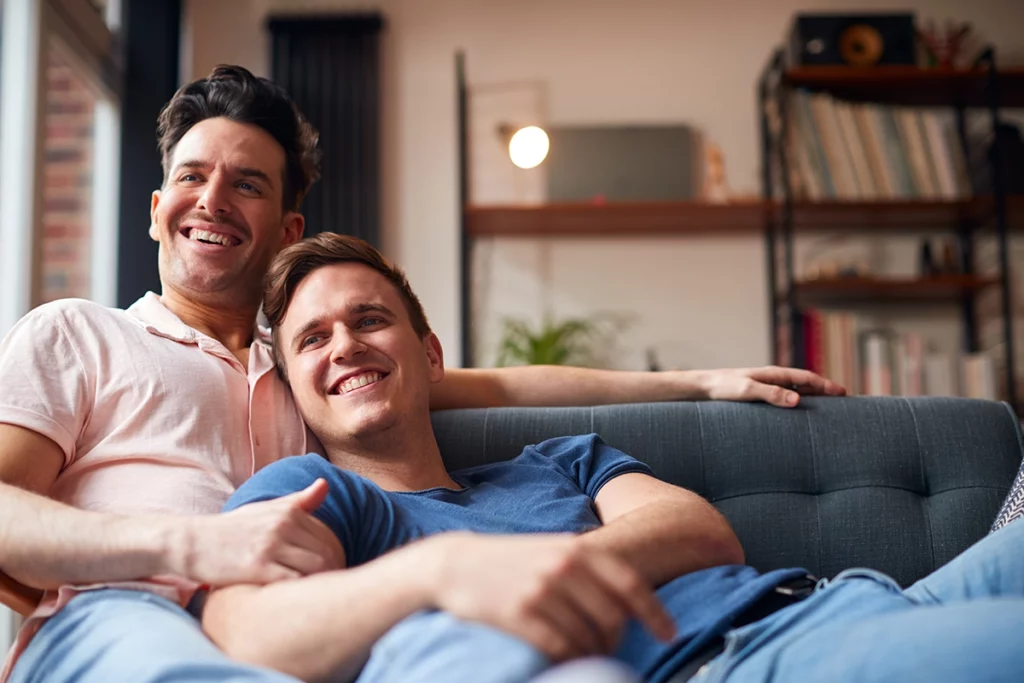 Loving Same Sex Male Couple Lying On Sofa At Home Watching TV And Relaxing Together