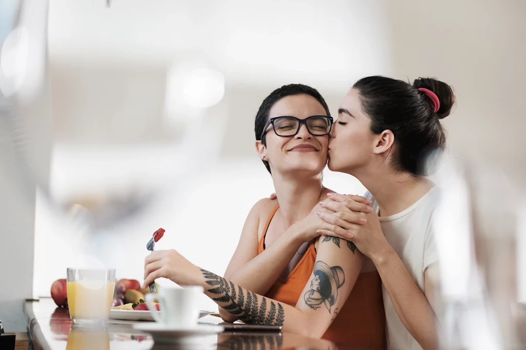 BlahFace.com - The Topic is Rainbow Connection, LGBT. A couple of gay woman having breakfast together and holding hands while smiling and kissing. Same sex young married female couple in their daily routine at a morning showing some affection LGBT